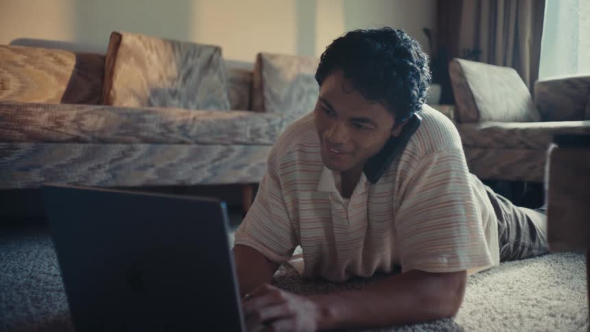 Man lying on carpet ending a call and interacting with a laptop in front of him