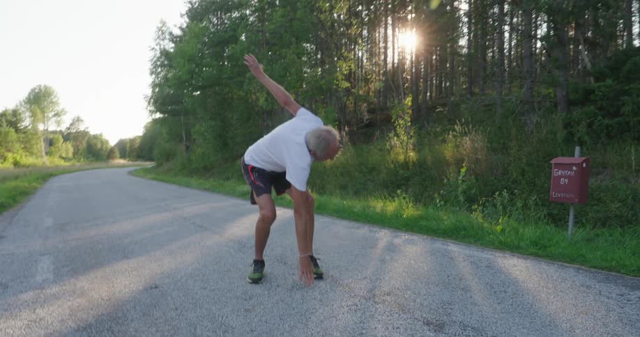 An elderly man stretches in the middle of the road next to a wooded area