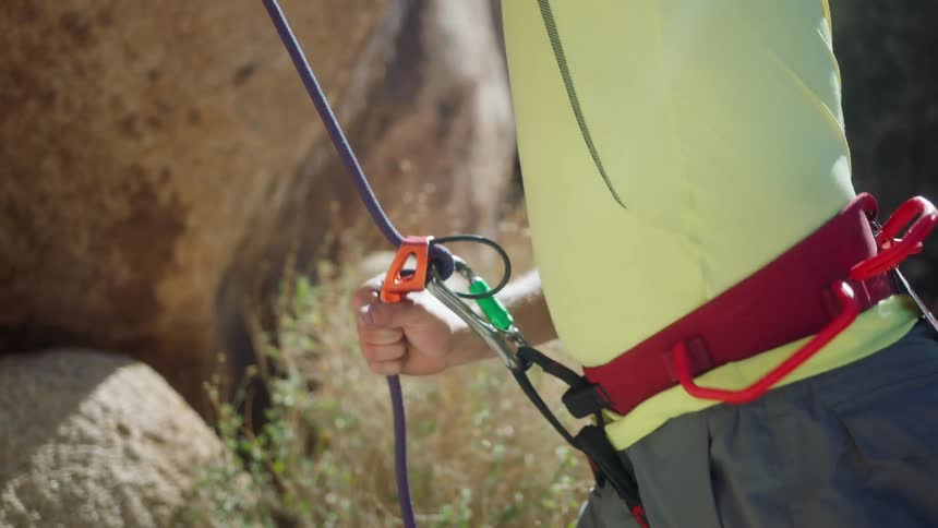 A closeup of a persons torso with climbing gear focusing on the belay device attached to the harness securing a rope