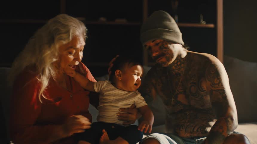 A tattooed man and an elderly woman sit on a couch the woman holding his baby but the baby is crying and restless