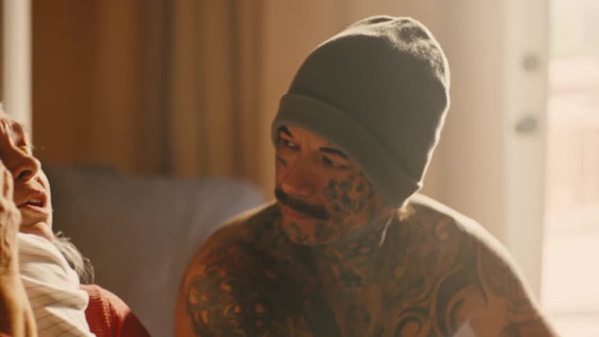 A man covered in tattoos wearing a snow cap sitting next to a woman holding a baby as they nod and converse