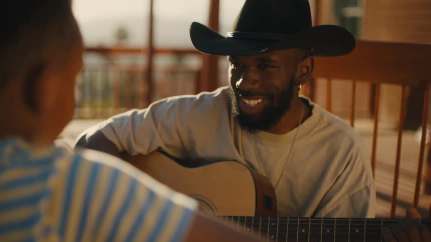 A father holds a guitar sitting in a rocking chair on a porch in front of his son as they laugh and smile