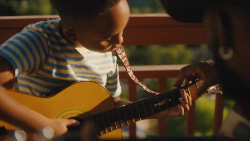 A father teaches his son to play guitar outdoors on a sunny day