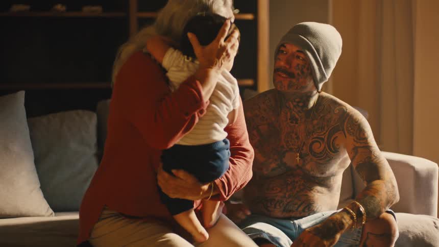A man holding a crying infant before handing the child to a woman sitting next to him on the couch as she rocks the child back and forth