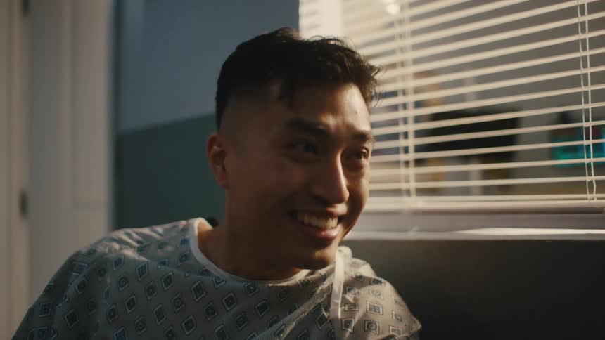 A man in hospital gown is sitting in an armchair talking to a nurse who is next to him and he is happy