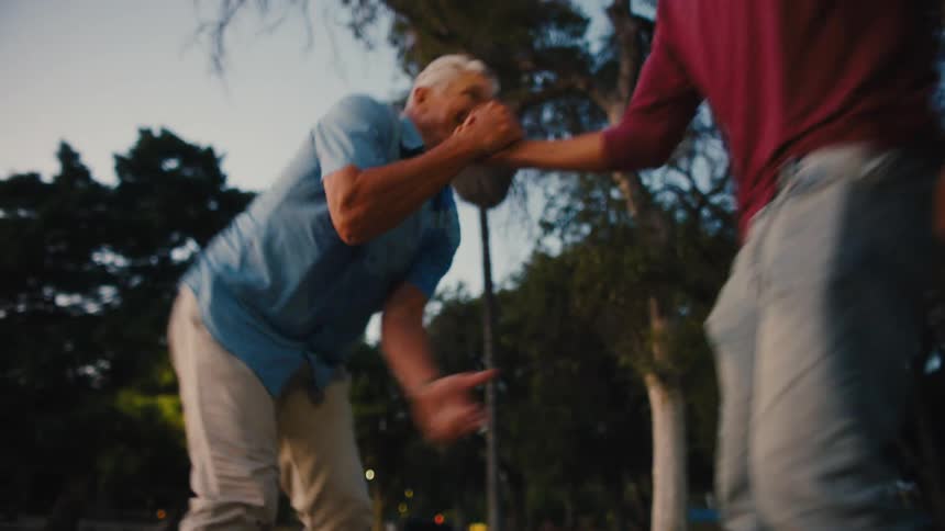A young man helps an elderly man stand up after he fell off a skateboard in a park