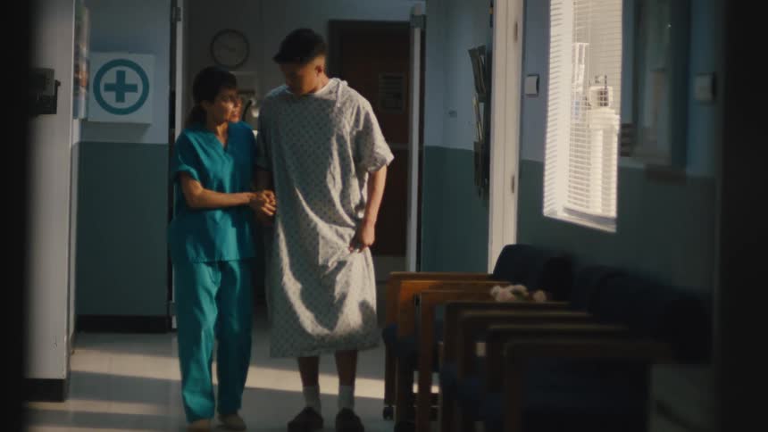 A man is walking with a limp with the help of a doctor who is accompanying him down the hallway