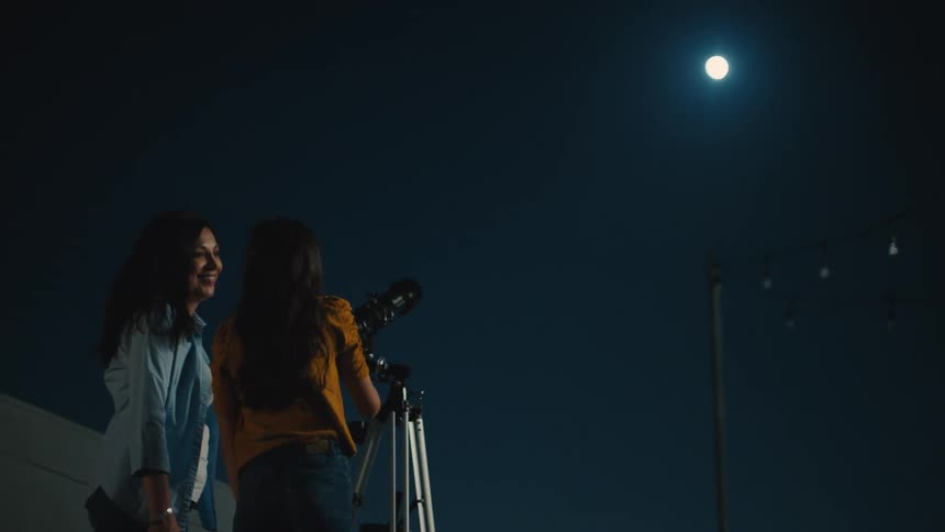 A mother and daughter looking through a telescope at the moon at night