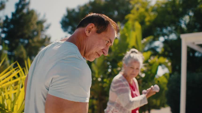 Man training biceps with dumbbell and in the background an elderly lady with smaller dumbbell repeating