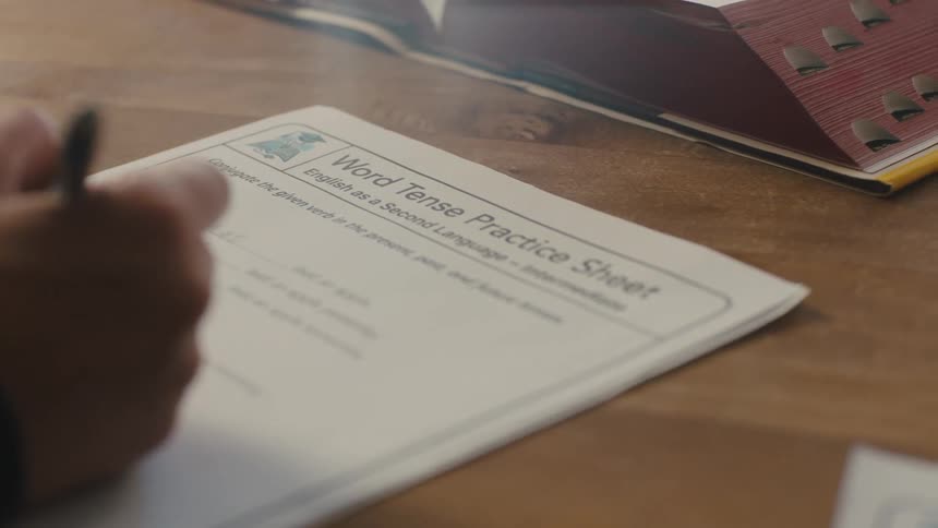 Close up of a white paper on a wooden table next to a large book with a persons hands writing on the paper with a pen
