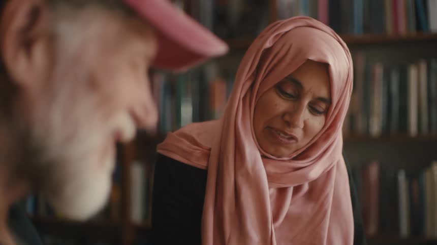 A woman wearing a pink scarf is looking down and smiling as she talks to a whitebearded man standing in front of her