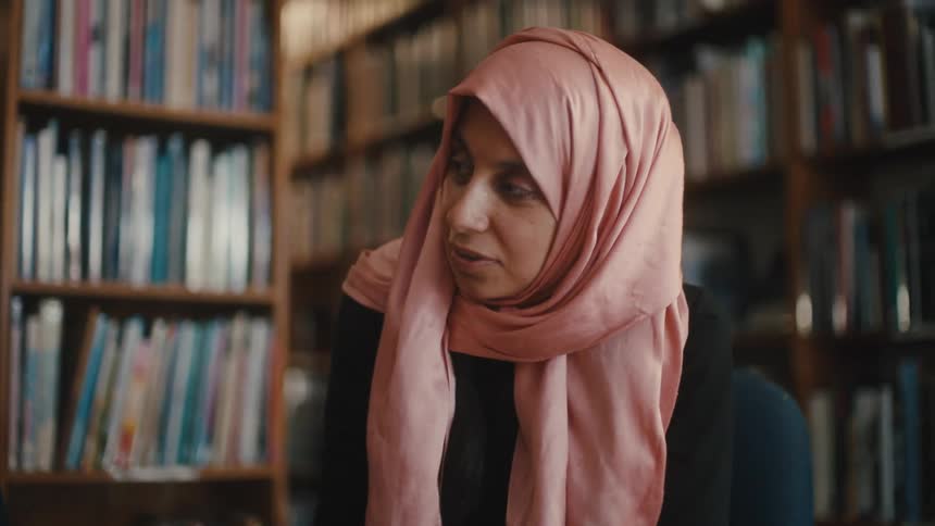 A bearded man explains a document to a woman wearing a pink headscarf in a library
