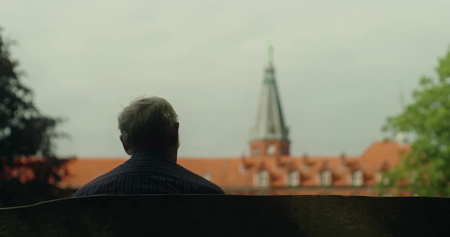 Back view of an elderly man sitting on a bench facing a classical building with a tower