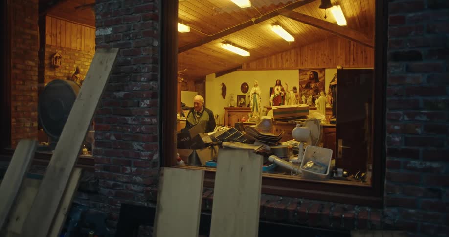 An older man walking through a building with lights on and odd items stacked everywhere