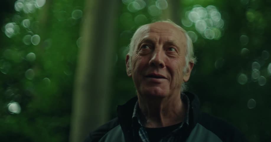 Close up of an older mans face as he walks through a forest with tall trees surrounding him