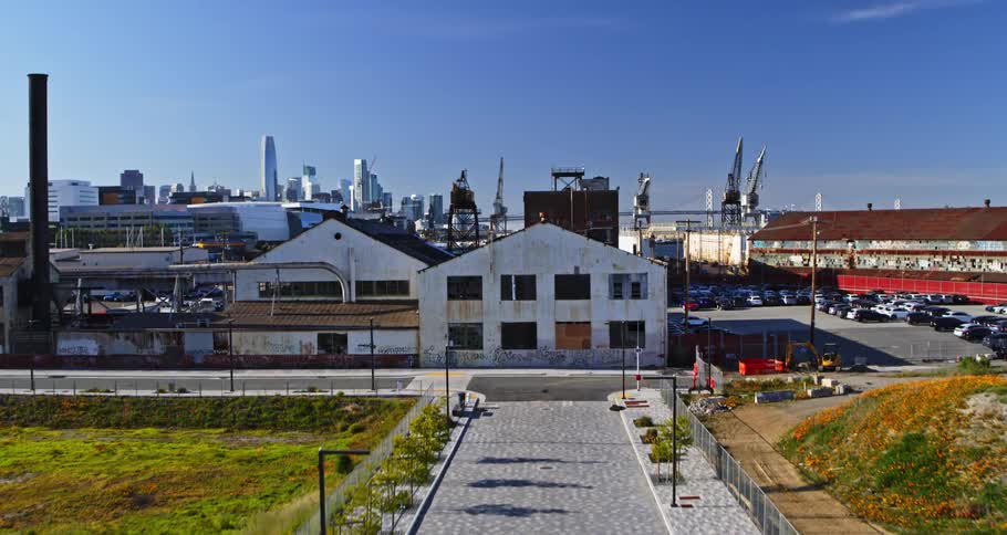 A concrete path with some trees on the sides that leads to a port with cranes and a parking lot with several cars next to a body of water