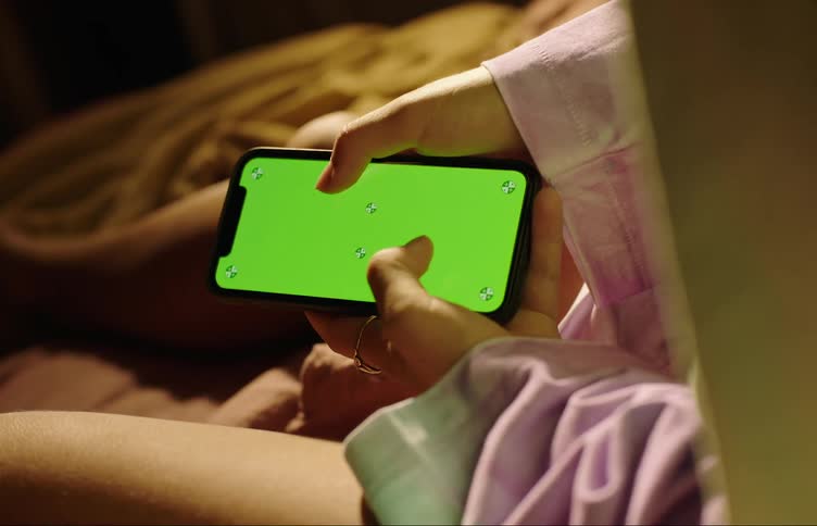 A person is using a smartphone with a green screen while sitting in bed