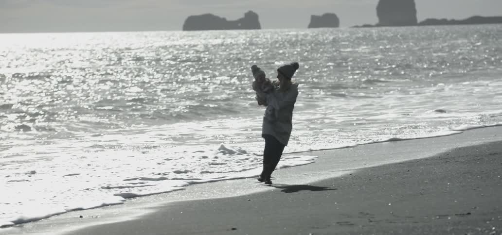 A woman carries a baby towards the ocean water and runs away playfully