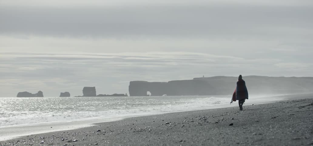 A woman wrapped in a blanket walks on a beach