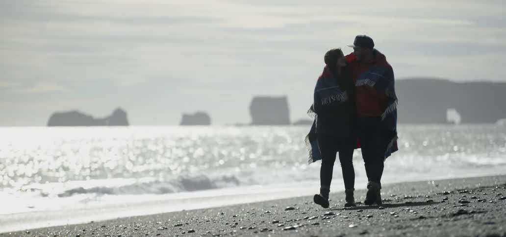 Couple walking on a cold beach sharing a blanket around their torsos on a cloudy day