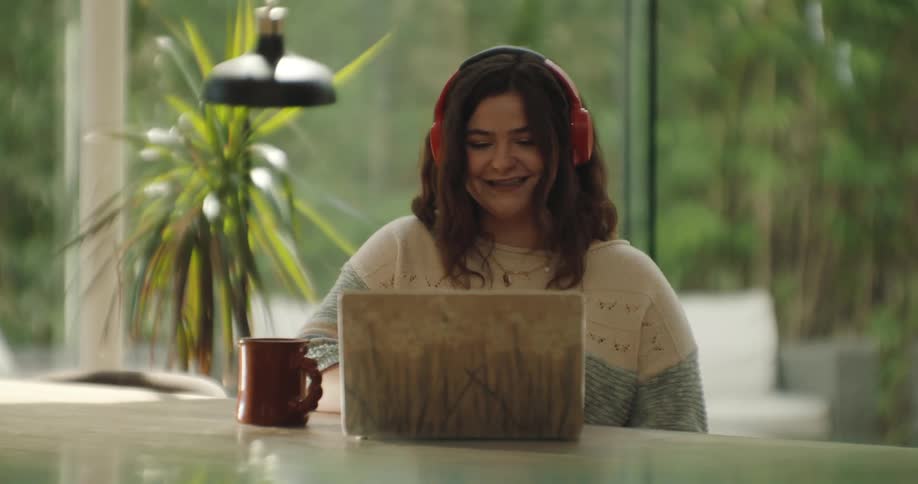 A woman sitting at a table on her laptop with headphones in and a mug next to her