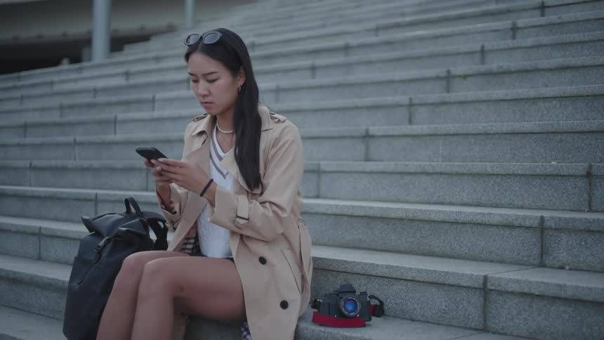 An asian woman sitting on concrete steps reads something on her smartphone