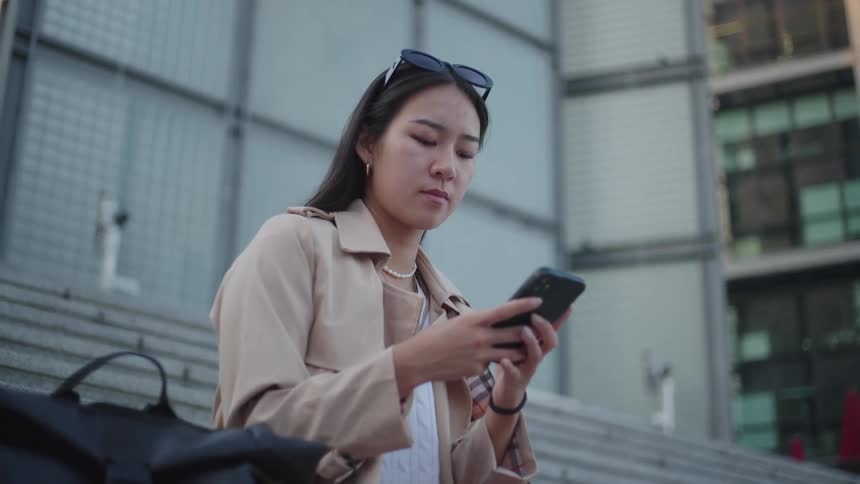 An asian woman sits on the steps outside of a city building and reads something on her smartphone