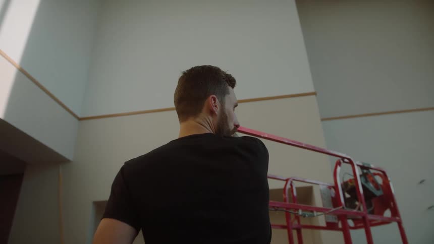 A man in a black shirt stands with his back turned and pointing to a white wall with a red metal structure in front