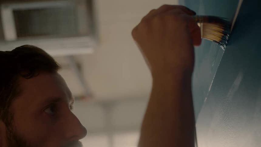 Closeup of a persons arm holding a paintbrush against a wall applying blue paint