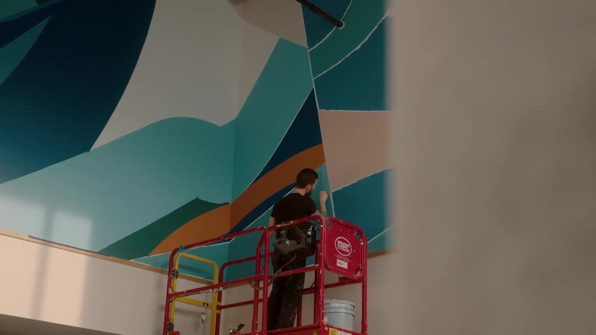 A man on top of a red structure who is using a paintbrush to paint a wall blue