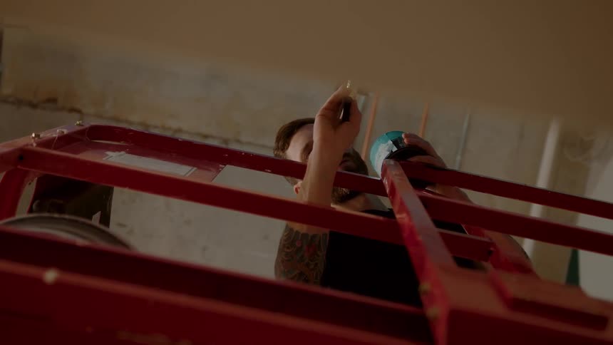 A man with a tattoo on his arm and is using a brush to paint a wall and he is standing on a red iron structure