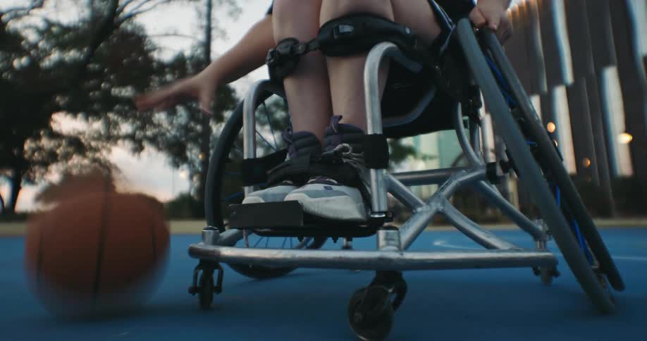 A person strapped into a wheelchair dribbles a basketball on a city court in a park