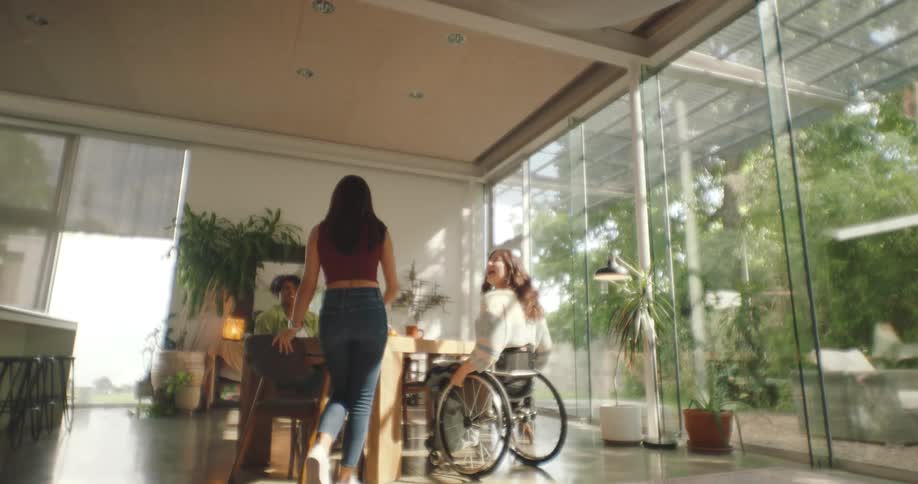 A group of friends sit at a wooden table one in a wheelchair near large windows looking at a patio
