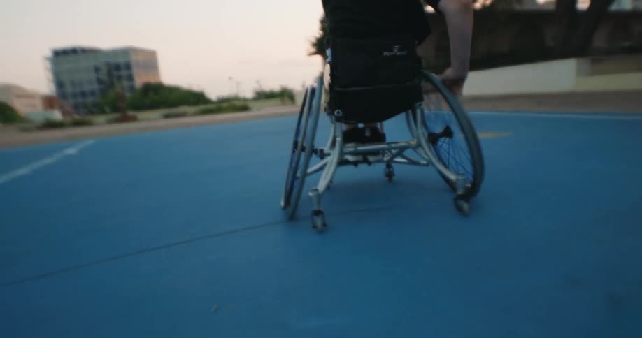 A woman in a wheelchair bouncing a basketball on the ground and making a basket and the court she is walking on is painted blue