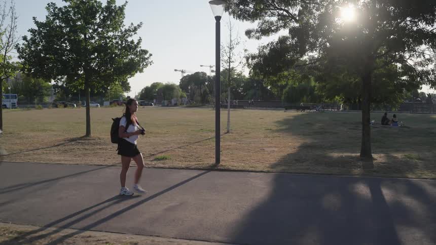 A young woman with a backpack is walking on a concrete path in an area with a large lawn and trees