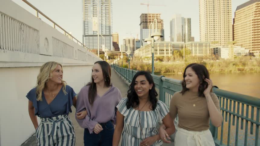 Four women walk down a walkway by a lake in a city