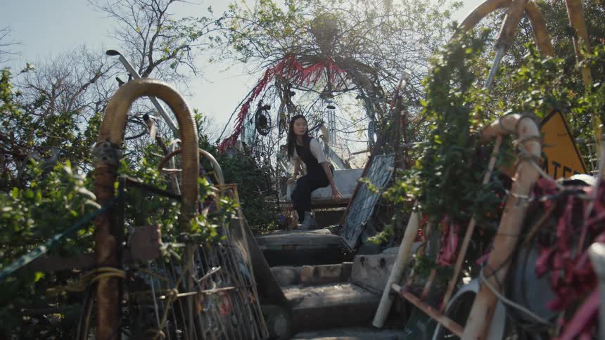 A young woman sits on a bench surrounded by structures made with cluttered objects