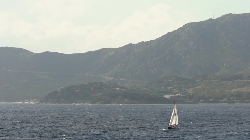 A single sailboat on choppy water with a mountainous coastline in the background under an overcast sky