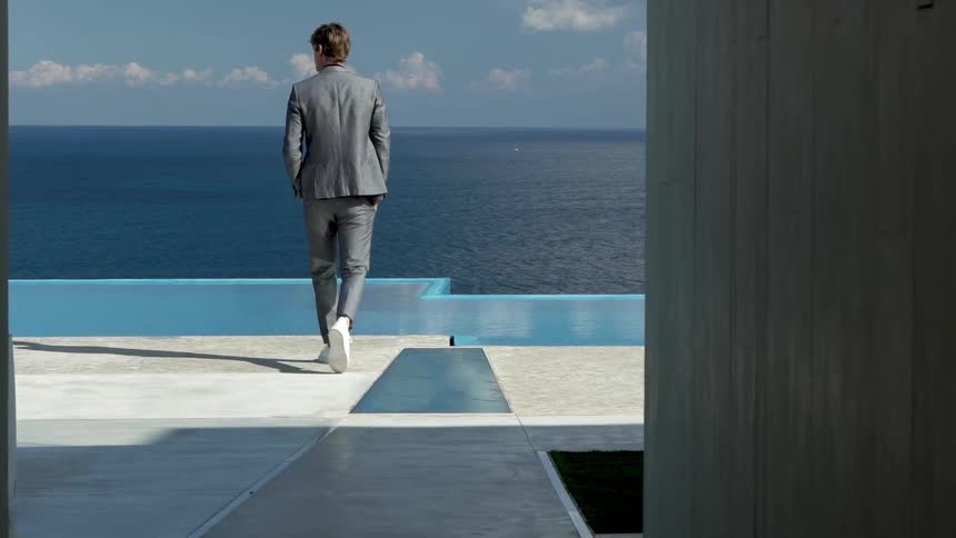 A man is standing with his back turned and wearing formal clothes and is walking in an outdoor setting to look at the ocean on the horizon