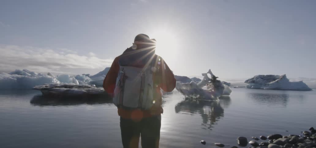 A man with a backpack walks to the water's edge and looks at icebergs