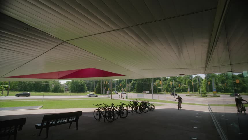 Bicycles parked under a pavilion near benches as traffic drives by on the road and a person rides in and parks his bike