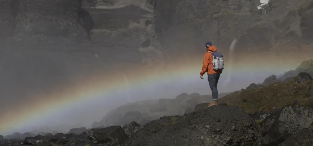 A hiker stands on a boulder overlooking a rainbow