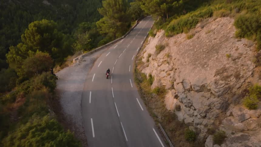 Motorcyclist drives along a highway through forested mountains passing a motorcyclist taking a break on the shoulder overlooking a forested slope
