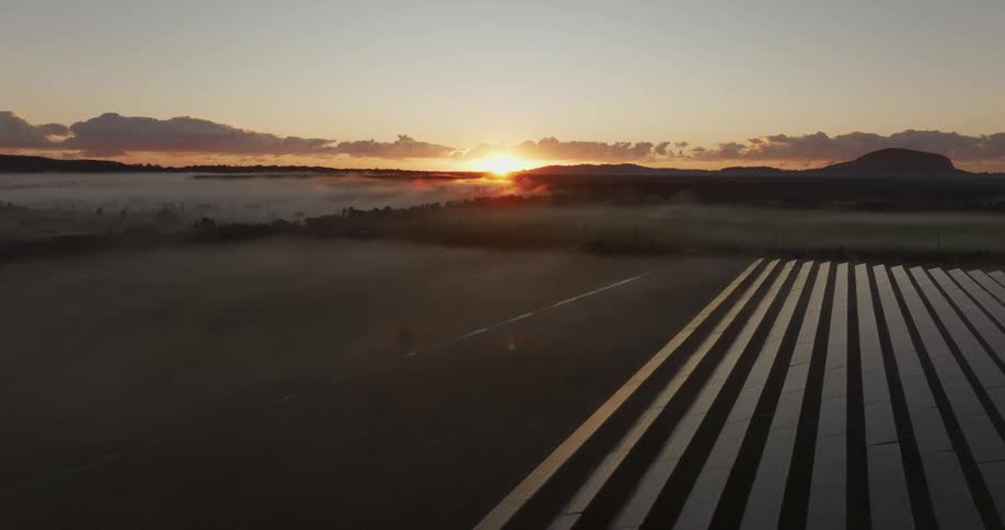 Aerial of solar panels in a wide open area with mountains in the distance and the sun setting on the horizon
