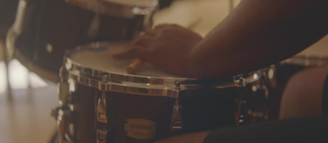 A man is playing drums using drumsticks and he is sitting while looking to his side and is wearing reading glasses