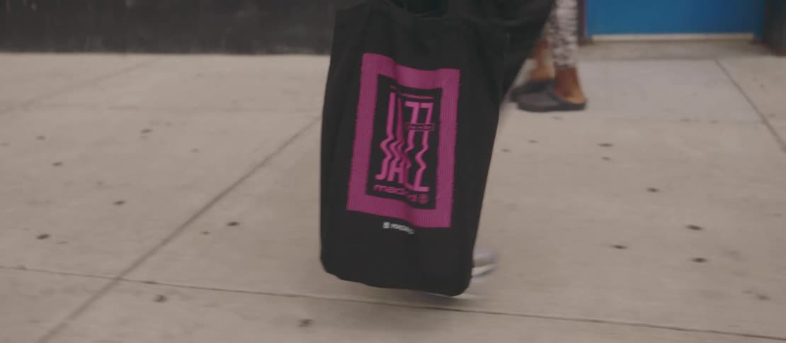 The legs of a person wearing white sneakers and carrying a black bag with pink details walking on a sidewalk