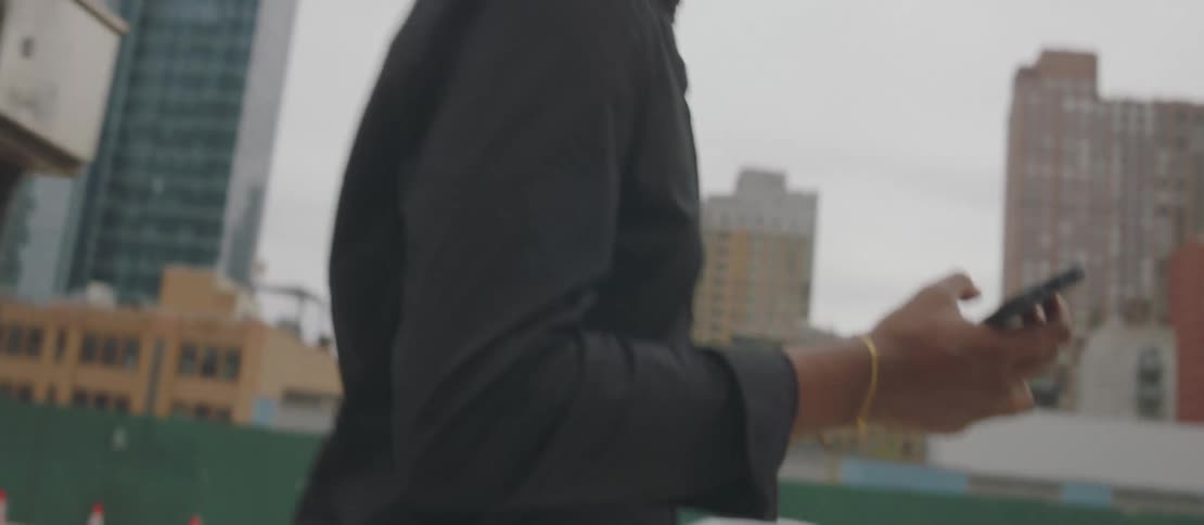 A young man wearing earbuds is walking looking at his cell phone that came out of his hand and going after him and there are several skyscraper buildings