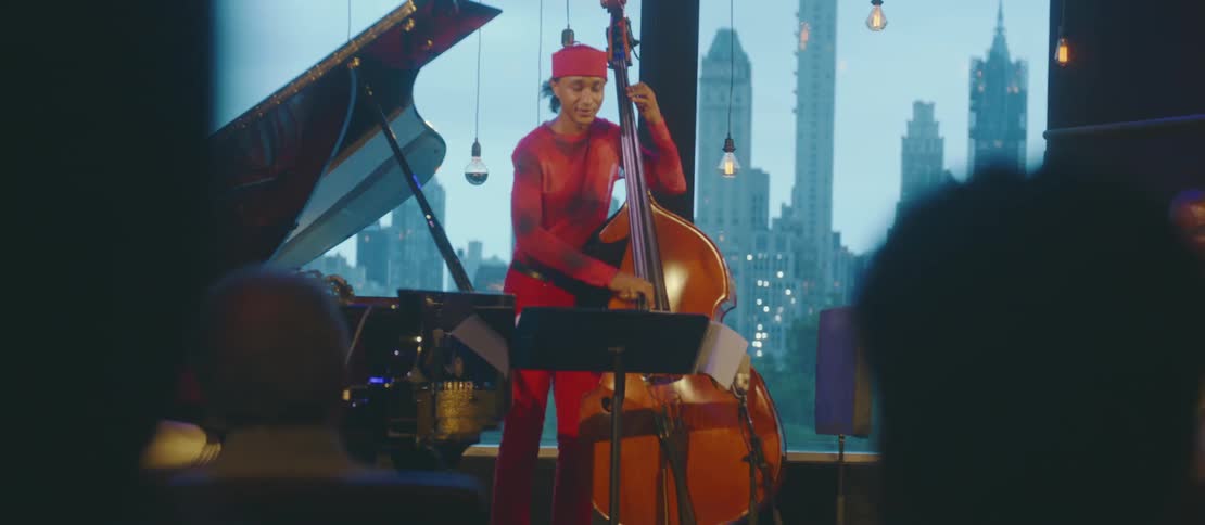 A man standing playing a violin celo next to a man playing the piano and another playing the drums and there are some people in front watching them