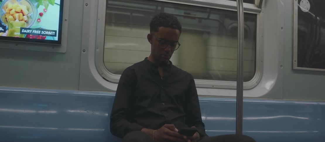 A young man sitting on a subway focuses on his smart phone as the train starts moving