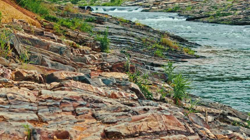 Rocky riverbank with stratified rocks and sparse green vegetation leading to a flowing river with small rapids